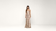A woman stands in a minimal studio wearing a sleeveless, fitted, patterned floor-length dress. She has long brown hair with bangs and is looking at the camera, with one hand resting by her side and the other on her hip.