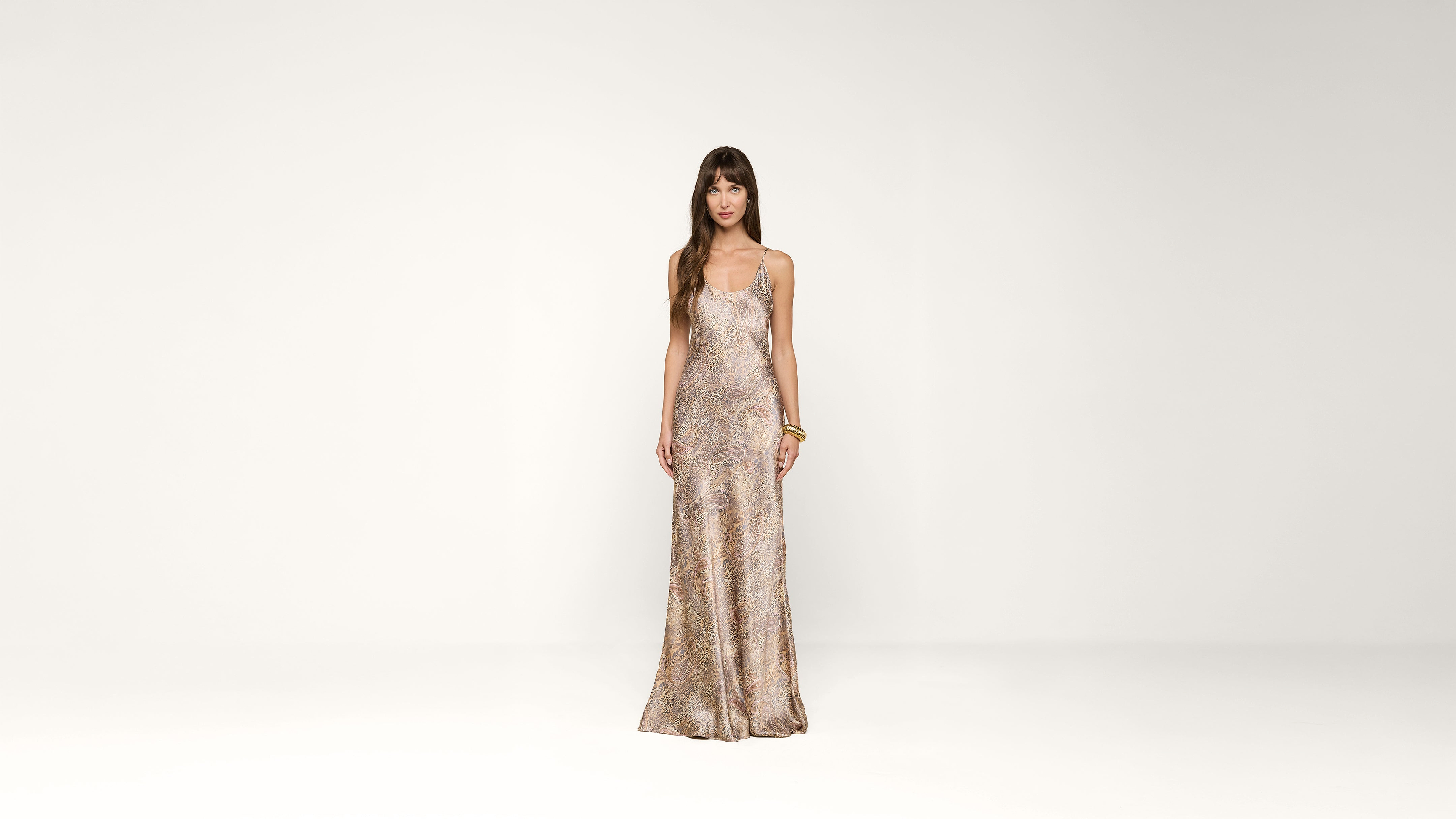 A woman stands in a minimal studio wearing a sleeveless, fitted, patterned floor-length dress. She has long brown hair with bangs and is looking at the camera, with one hand resting by her side and the other on her hip.