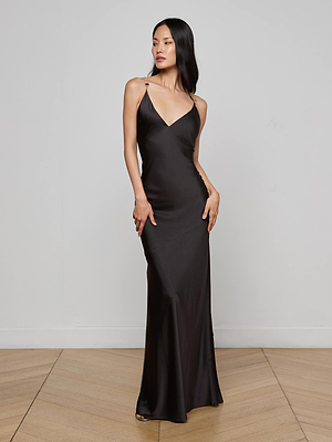 A woman stands on a wooden floor wearing the L'AGENCE Saint Crystal Strap Gown, a long, dark brown satin dress with thin straps, posing with one hand on her hip against a plain light gray wall.