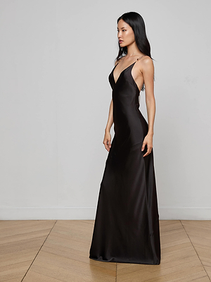 A woman with long black hair stands sideways against a white wall and wooden floor, wearing the L'AGENCE Saint Crystal Strap Gown—a sleeveless, floor-length black satin gown with crystal straps and a deep V-neckline.
