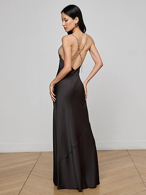 A woman with long dark hair stands in a minimalist room, wearing the L'AGENCE Saint Crystal Strap Gown—a floor-length, open-back satin dress with thin crisscross straps—posing with one hand on her hip and looking over her shoulder.