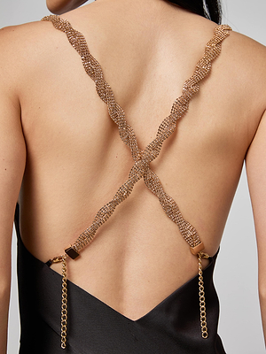 A close-up of a woman's back wearing the L'AGENCE Saint Crystal Strap Gown, featuring an open back with crisscrossed gold mesh straps and a delicate gold chain draping at the lower back.