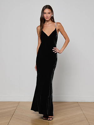 A woman poses confidently with one hand on her hip, wearing the L'AGENCE Saint Velvet Gown. The elegant black dress features crystal-embellished straps, a deep V-neckline, and an open back. She has long, straight hair and stands against a plain wall.