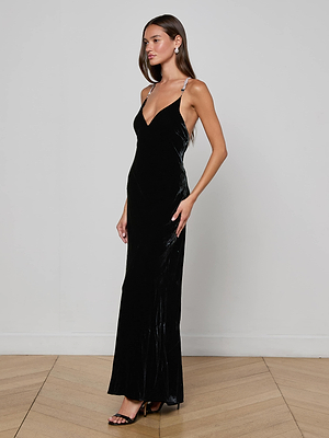 A woman stands in profile against a white wall and wooden floor, wearing the L'AGENCE Saint Velvet Gown—a long black velvet dress with a deep V-neck and crystal straps—paired with black high-heeled sandals.