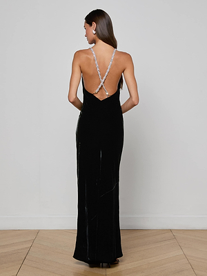 A woman faces away wearing the L'AGENCE Saint Velvet Gown, a long black velvet dress with an open back and crisscrossed crystal straps, standing on a wooden floor against a plain white wall.