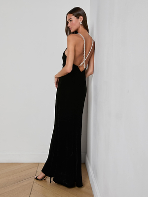 A woman with straight brown hair and minimal accessories faces away from a white wall, wearing the L'AGENCE Saint Velvet Gown—a long black, crystal-strap dress with an open back—paired with black high heels.