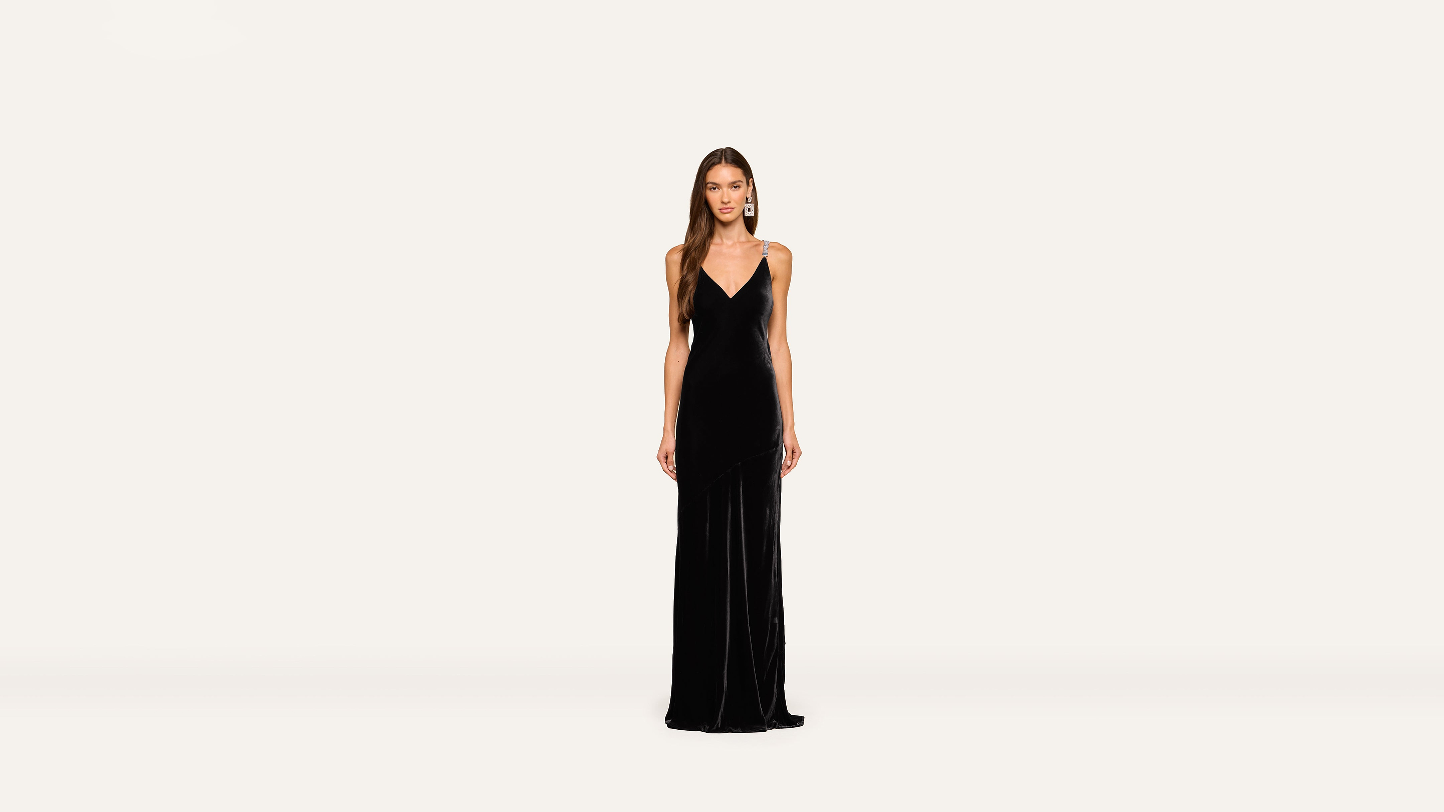 A woman stands facing forward in a long, sleeveless black gown with a V-neckline against a plain white background, exuding elegance and poise.