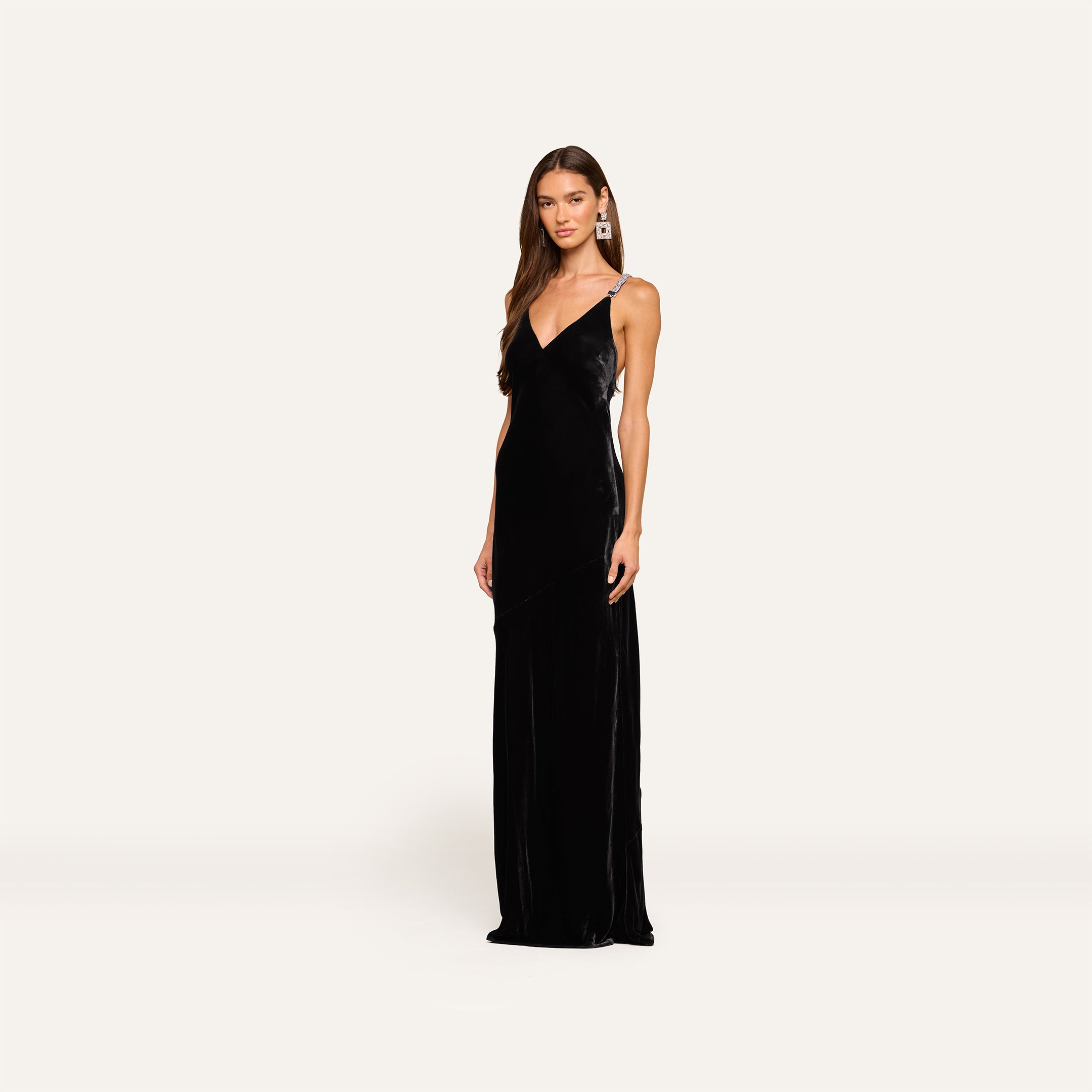 Woman in the Saint Velvet Gown—a long black velvet dress with crystal and thin straps—stands against a light background, her long brown hair styled and large statement earrings on.