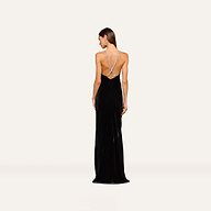 A woman faces away, wearing the Saint Velvet Gown—a long black velvet dress with a low open back and crisscross crystal straps—against a plain light background.