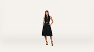 A woman in a sleeveless black dress with a belt stands confidently with hands in her pockets, wearing black heels against a plain white background.
