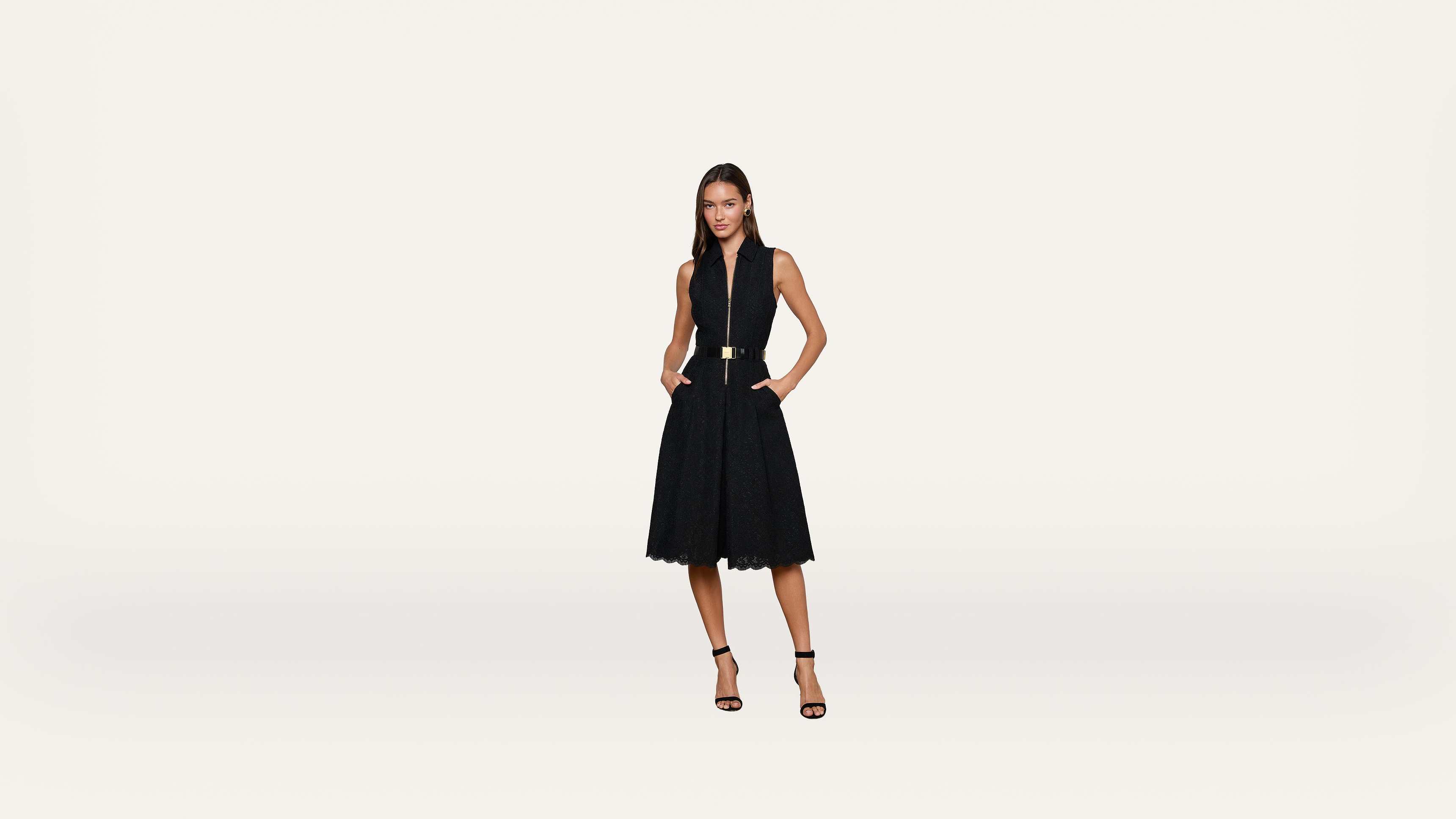 A woman in a sleeveless black dress with a belt stands confidently with hands in her pockets, wearing black heels against a plain white background.