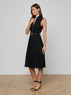 A woman poses indoors wearing the Kylo Lace Shirt Dress, featuring a sleeveless black floral lace design with zip front and belted waist. She stands on a wooden floor against a white wall, accessorized with black heels and gold earrings.