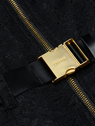 Close-up of a gold metal buckle with LAGENCE engraved, attached to a black strap and zippered, textured black fabric—an elegant detail that elevates the Kylo Lace Shirt Dress, perfect for adding polish to your favorite pocketed look or taking your style seamlessly from desk to drinks.