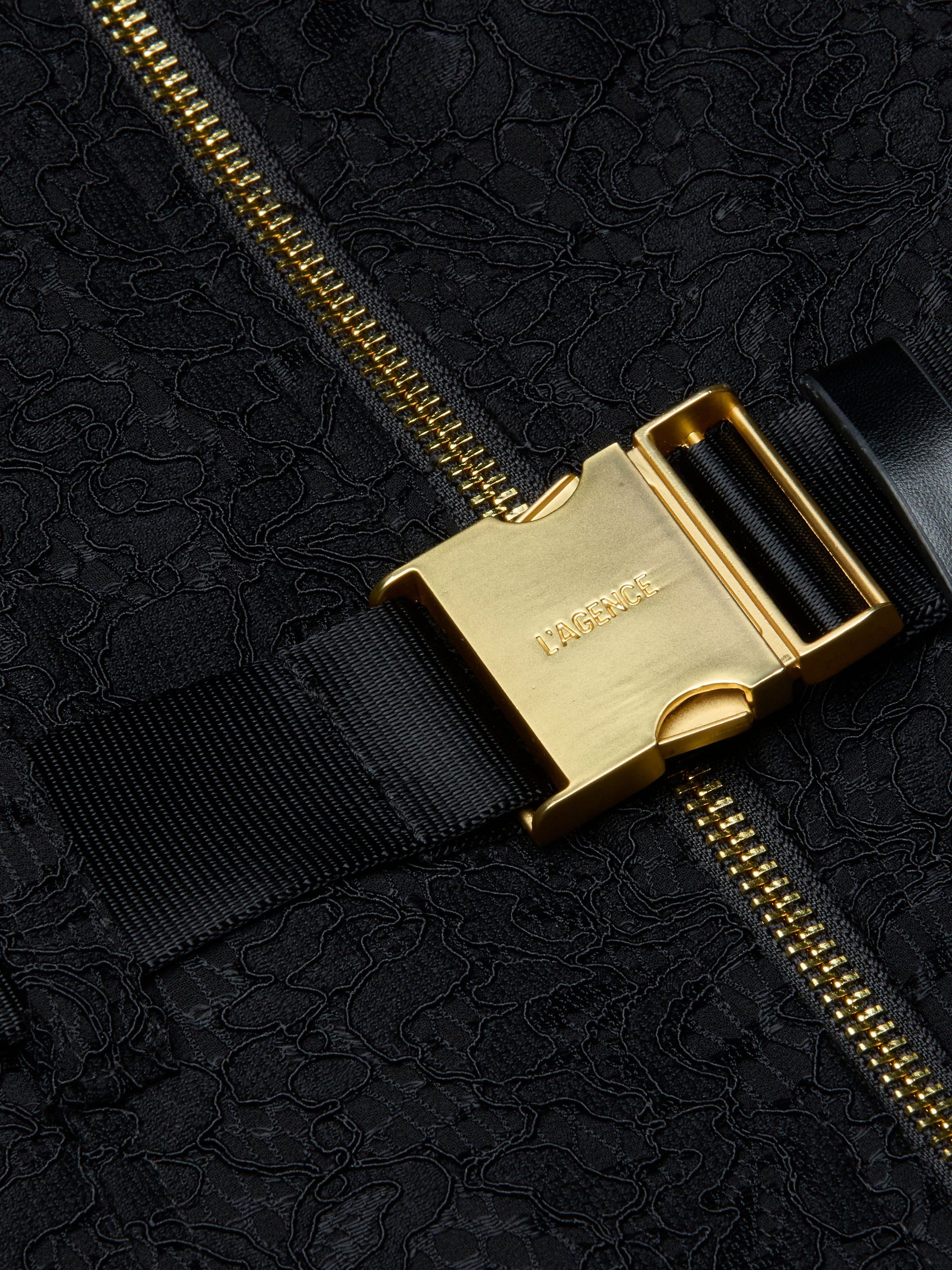 Close-up of a gold metal buckle with LAGENCE engraved, attached to a black strap and zippered, textured black fabric—an elegant detail that elevates the Kylo Lace Shirt Dress, perfect for adding polish to your favorite pocketed look or taking your style seamlessly from desk to drinks.