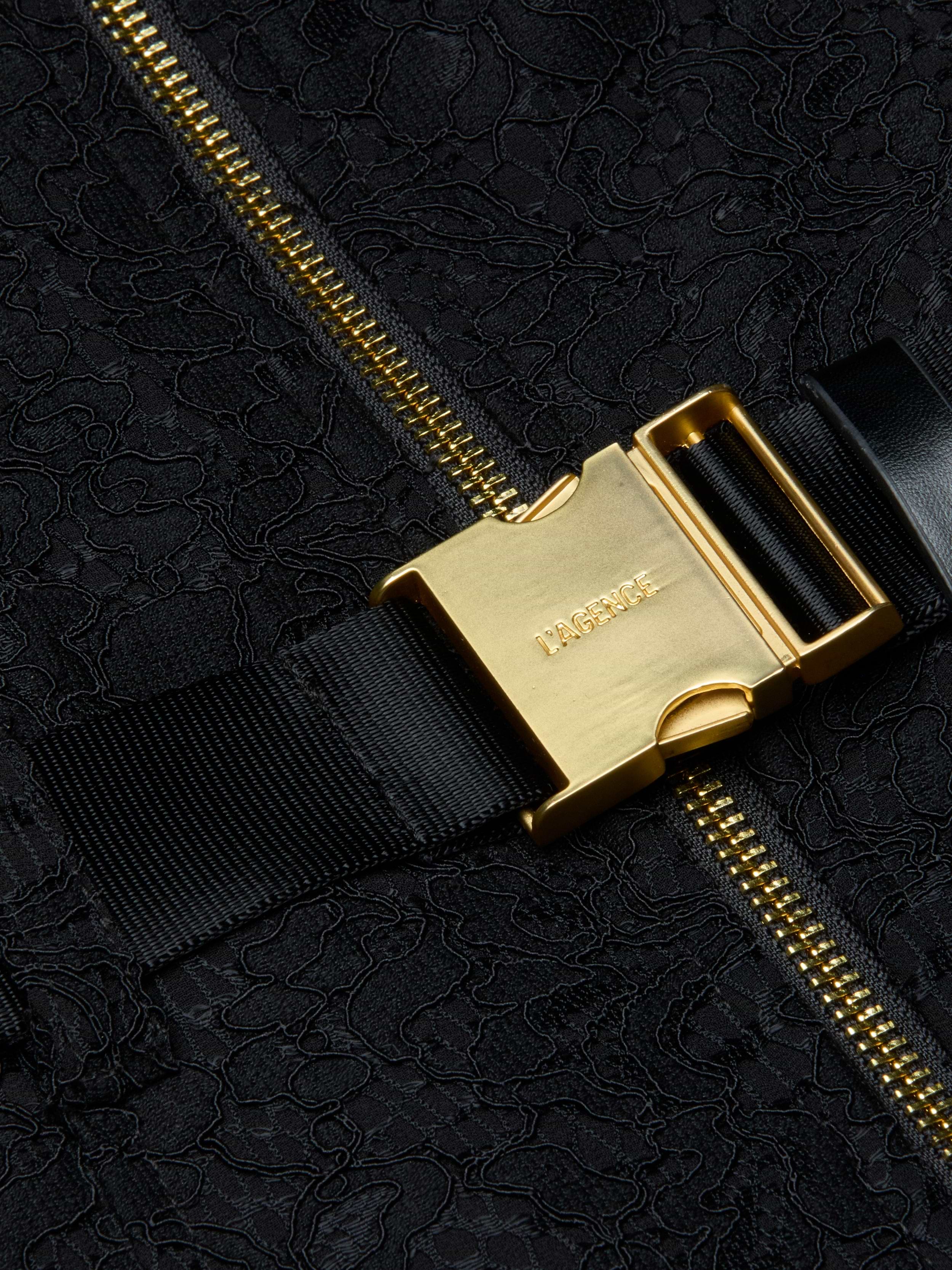 Close-up of a gold metal buckle with LAGENCE engraved, attached to a black strap and zippered, textured black fabric—an elegant detail that elevates the Kylo Lace Shirt Dress, perfect for adding polish to your favorite pocketed look or taking your style seamlessly from desk to drinks.