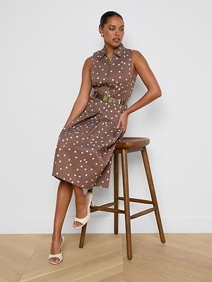 Woman wearing a brown polka dot dress on a wooden stool against a white wall.