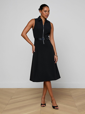 A woman poses confidently in a minimal room wearing the Kylo Shirt Dress, a black structured suit dress with a belted waist, paired with black high-heeled sandals. She stands with one hand on her hip and looks to the side.