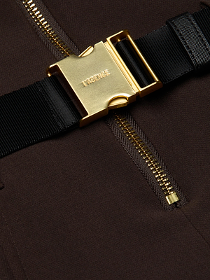 Close-up of the Kylo Shirt Dress by L’AGENCE: a black fabric strap with a shiny gold buckle fastened over dark brown suit fabric, featuring a prominent gold zipper running vertically beneath the strap.