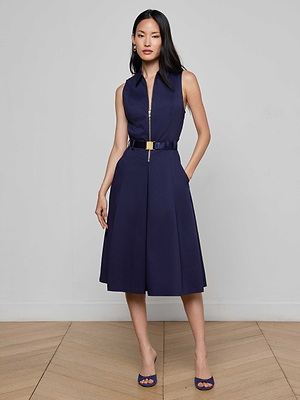 Indoors on a wooden floor, a woman wears the Kylo Poplin Shirt Dress—a sleeveless navy cotton dress with a front zipper, belted waist, and pockets—styled with navy heels and gold hoop earrings for a desk-to-drinks look.