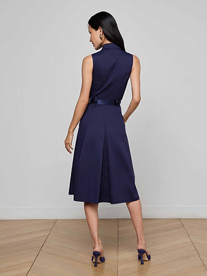 A woman stands on a wooden floor facing away, wearing the Kylo Poplin Shirt Dress in sleeveless dark blue cotton with pockets and a belt, paired with matching high-heeled shoes. The background is a plain light gray wall.