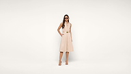 A woman wearing a sleeveless beige dress, sandals, and sunglasses stands confidently against a plain white background with her hands in her pockets.