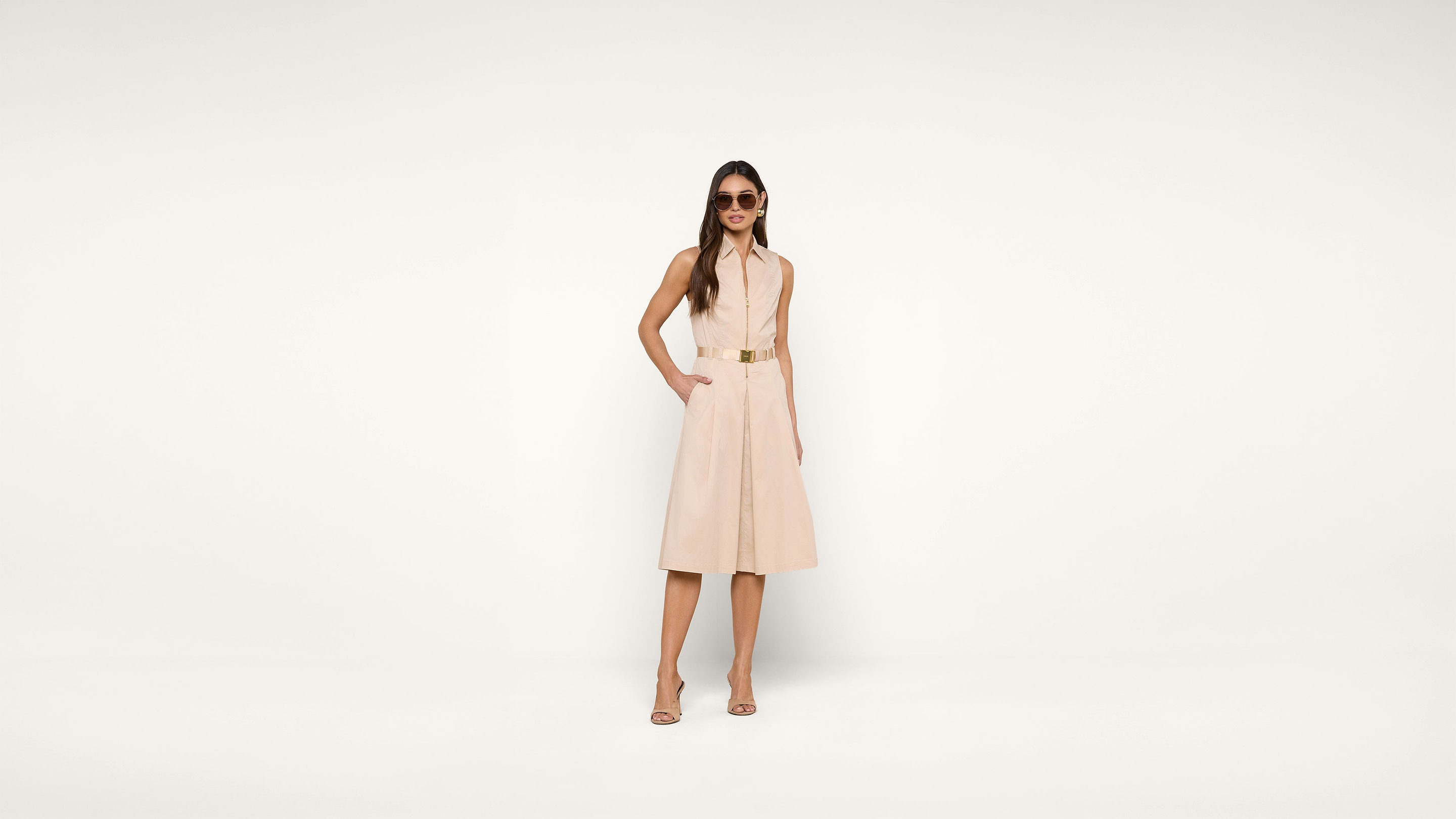 A woman wearing a sleeveless beige dress, sandals, and sunglasses stands confidently against a plain white background with her hands in her pockets.