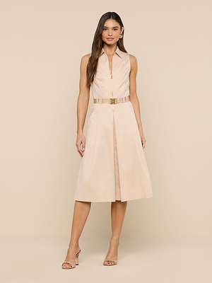 A woman stands against a beige background wearing the Kylo Poplin Shirt Dress—a sleeveless, knee-length belted dress with a zippered front and pleated skirt. She accessorizes with gold earrings, open-toed heels, and has long hair.