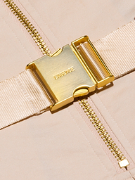 Close-up of a shiny gold buckle engraved with LAGENCE, attached to a beige fabric strap—part of the Kylo Poplin Shirt Dress—lying across a gold zipper on a light beige background.