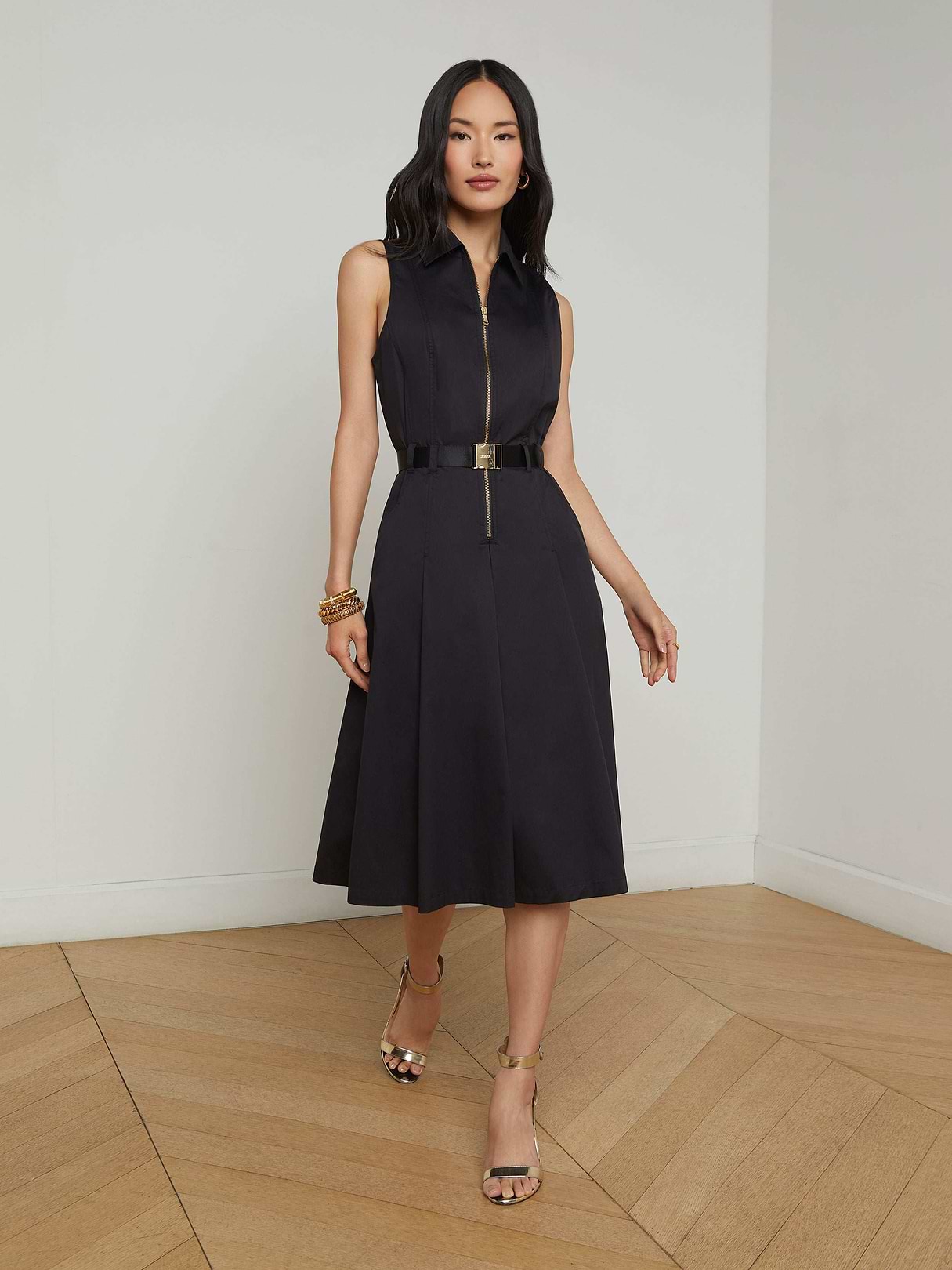 A woman in the Kylo Poplin Shirt Dress—a sleeveless black dress with a belt—stands against a plain white background, styled with gold heels and bracelets. Her hair is down, and her hands are relaxed at her sides. This versatile piece offers effortless style for any occasion, seamlessly taking you from desk to drinks.