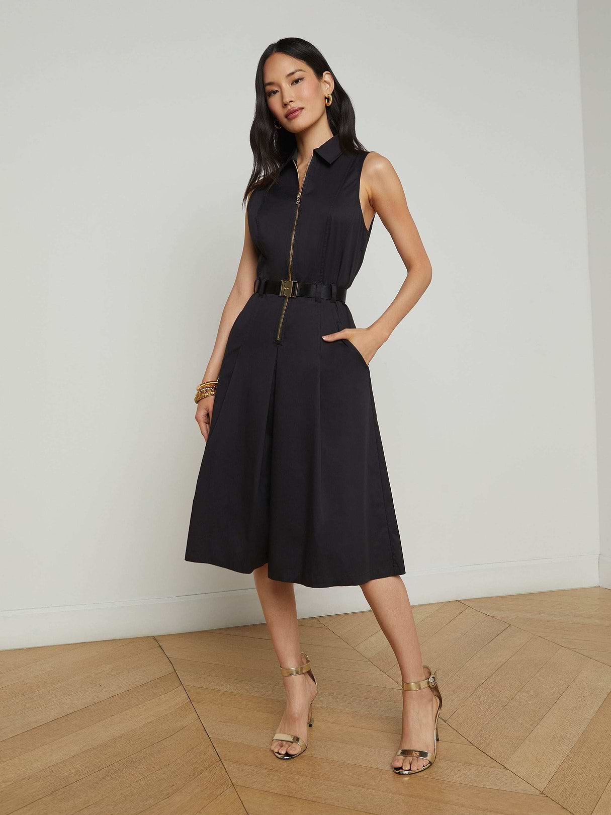 Woman wearing the Kylo Poplin Shirt Dress—a sleeveless, knee-length black dress featuring pockets and a belt—with her hands in the pockets, styled with strappy heels and gold jewelry, standing against a plain white background.