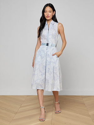 A woman poses indoors against a light background in the Kylo Shirt Dress—a sleeveless, light blue lace print midi with floral patterns and pockets—styled with silver high-heeled sandals for an ideal desk-to-drinks look.