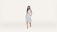 A woman stands on a plain white background, wearing a sleeveless light blue dress with a belt and sandals, with one hand in her pocket.