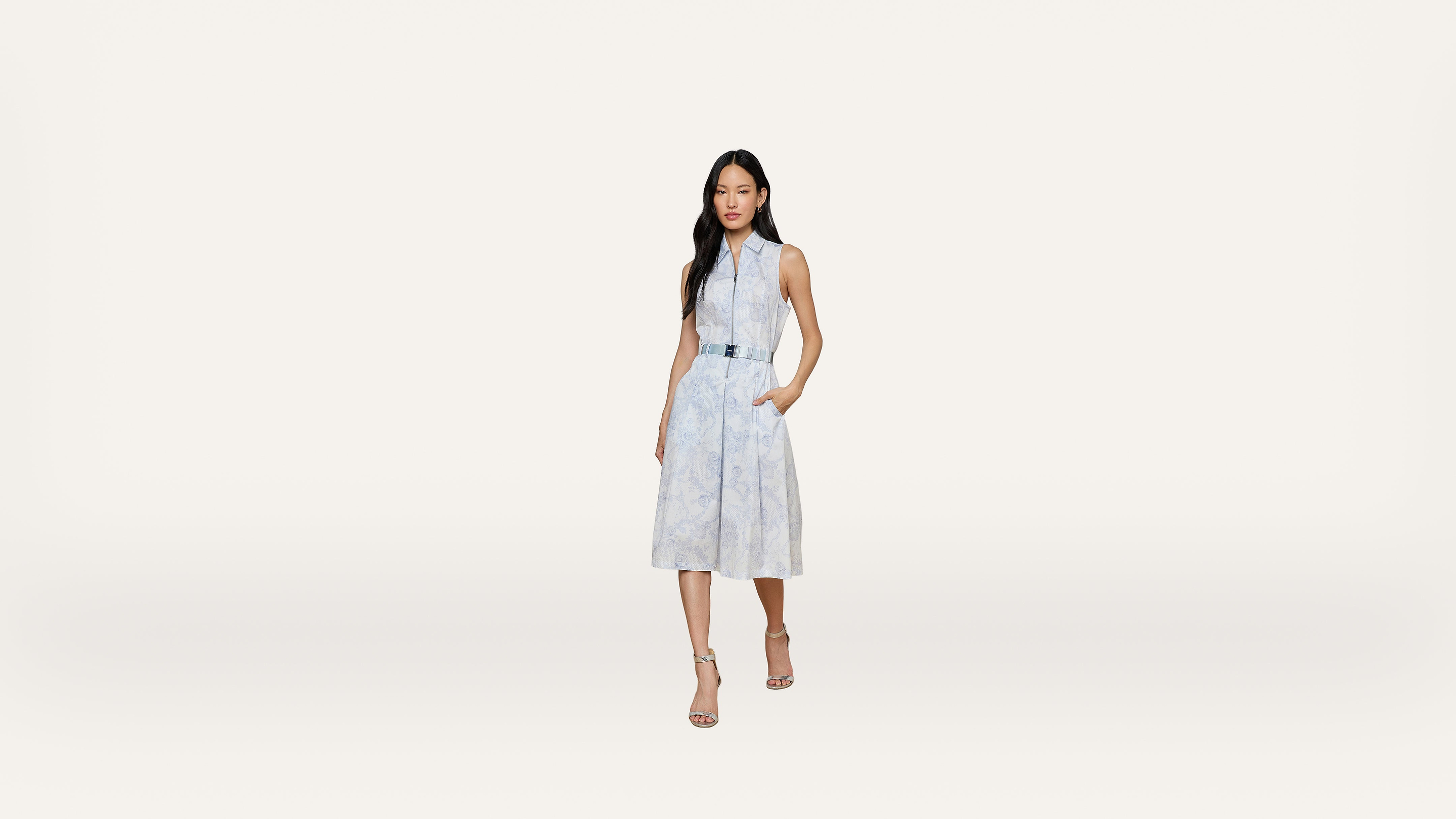 A woman stands on a plain white background, wearing a sleeveless light blue dress with a belt and sandals, with one hand in her pocket.