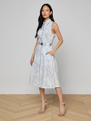 A woman with long dark hair stands on a wooden floor wearing the Kylo Shirt Dress—a sleeveless, collared, belted style with a subtle blue lace print—paired with silver high-heeled sandals for a perfect desk-to-drinks look.