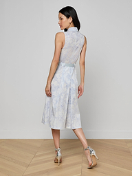 A woman stands on a light wooden floor facing away, wearing the Kylo Shirt Dress—a sleeveless blue lace midi shirt dress with a sheer lace back—paired with silver high-heeled sandals against a plain light wall for a chic desk-to-drinks look.