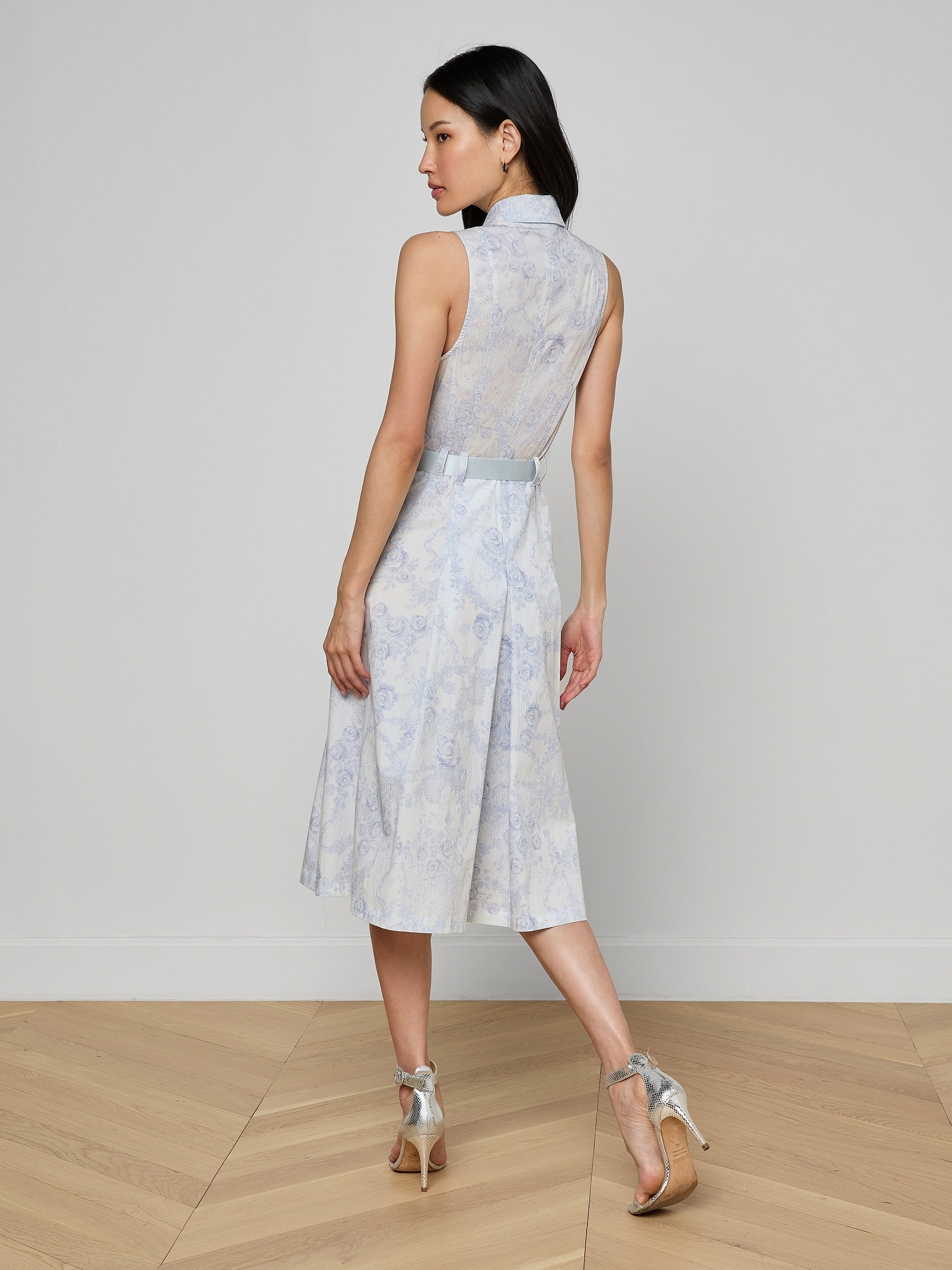A woman stands on a light wooden floor facing away, wearing the Kylo Shirt Dress—a sleeveless blue lace midi shirt dress with a sheer lace back—paired with silver high-heeled sandals against a plain light wall for a chic desk-to-drinks look.