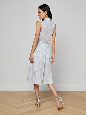 A woman stands on a light wooden floor facing away, wearing the Kylo Shirt Dress—a sleeveless blue lace midi shirt dress with a sheer lace back—paired with silver high-heeled sandals against a plain light wall for a chic desk-to-drinks look.