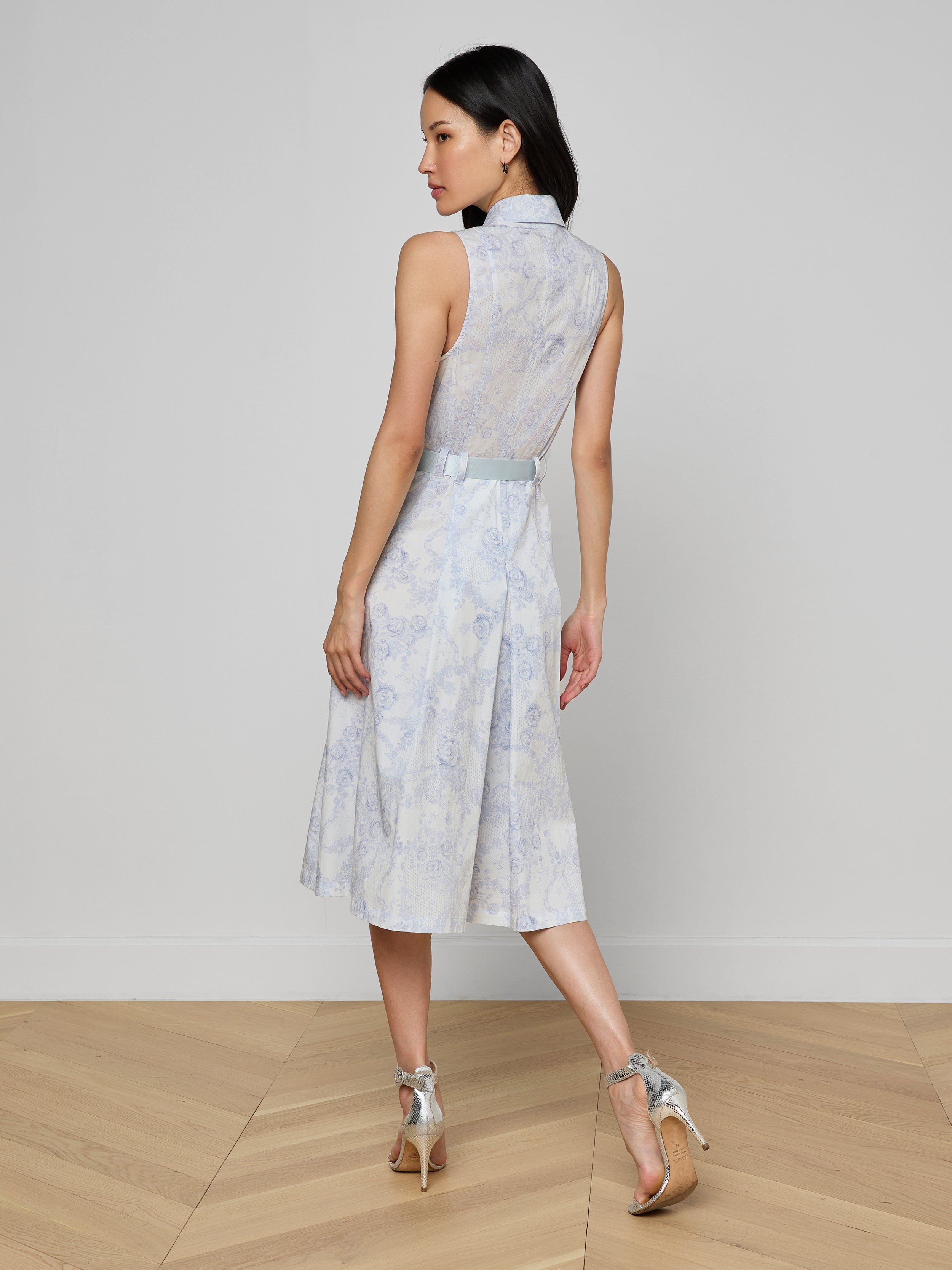 A woman stands on a light wooden floor facing away, wearing the Kylo Shirt Dress—a sleeveless blue lace midi shirt dress with a sheer lace back—paired with silver high-heeled sandals against a plain light wall for a chic desk-to-drinks look.