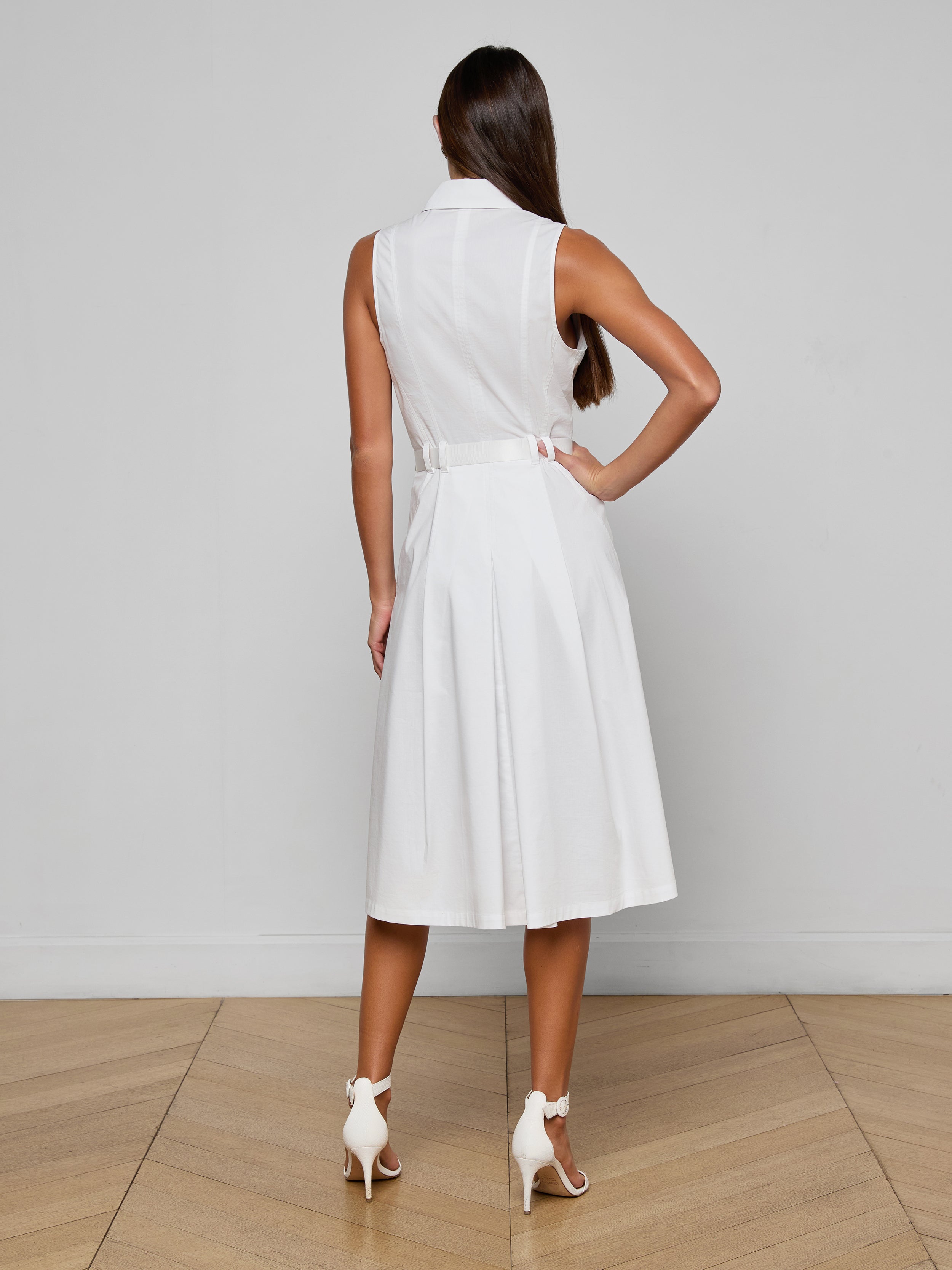 A woman with long brown hair wears the Kylo Poplin Shirt Dress—an elegant, sleeveless white dress made from organic cotton—and white high-heeled shoes, standing with her back to the camera against a plain white background.