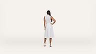 A woman with long dark hair stands facing away, wearing a sleeveless white dress and white heels, against a plain white background.