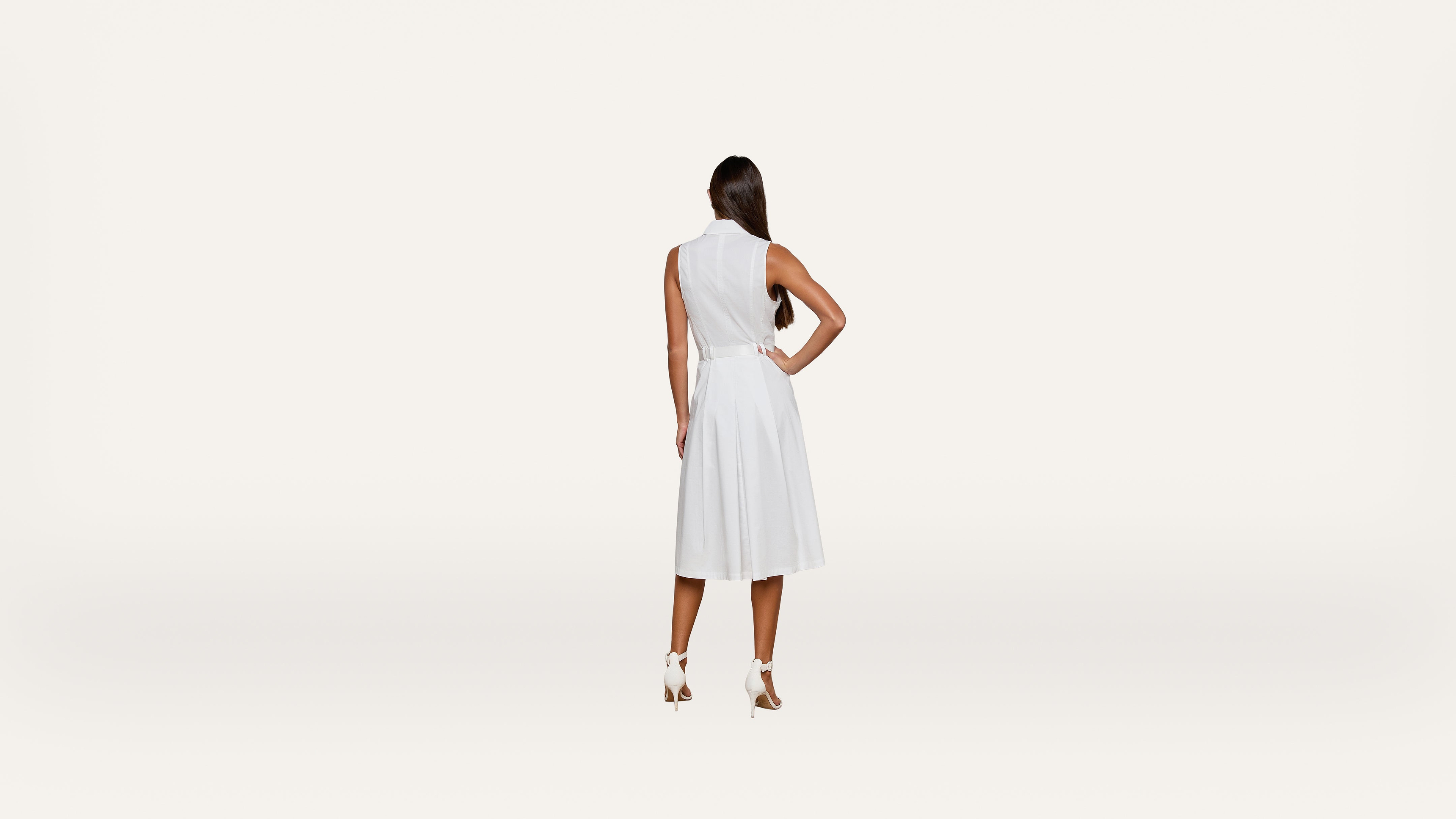 A woman with long dark hair stands facing away, wearing a sleeveless white dress and white heels, against a plain white background.