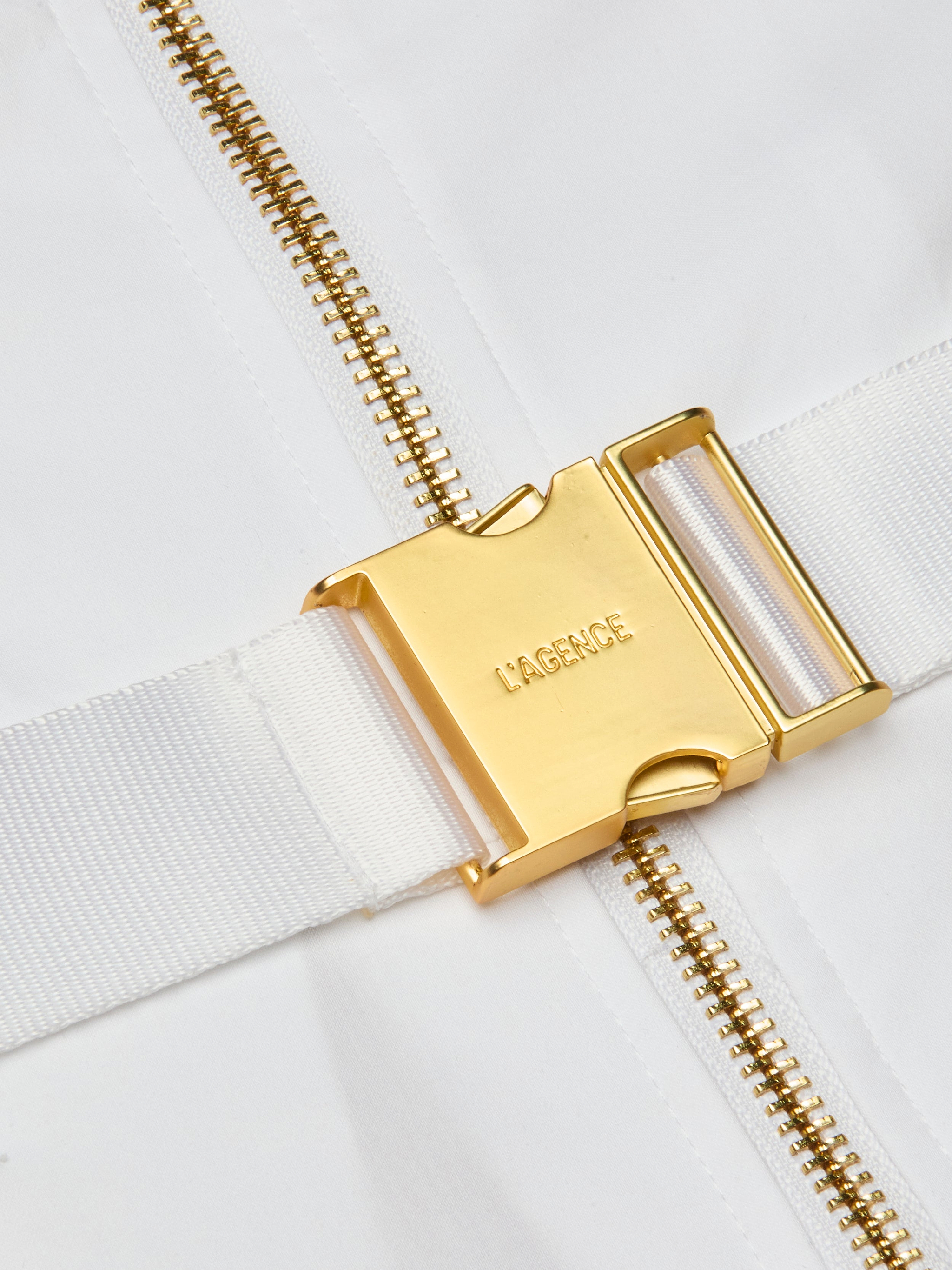 Close-up of a gold metal buckle engraved with LAGENCE on a white fabric strap, next to a gold zipper on a white background, highlighting the refined finish of the Kylo Poplin Shirt Dress crafted from organic cotton.