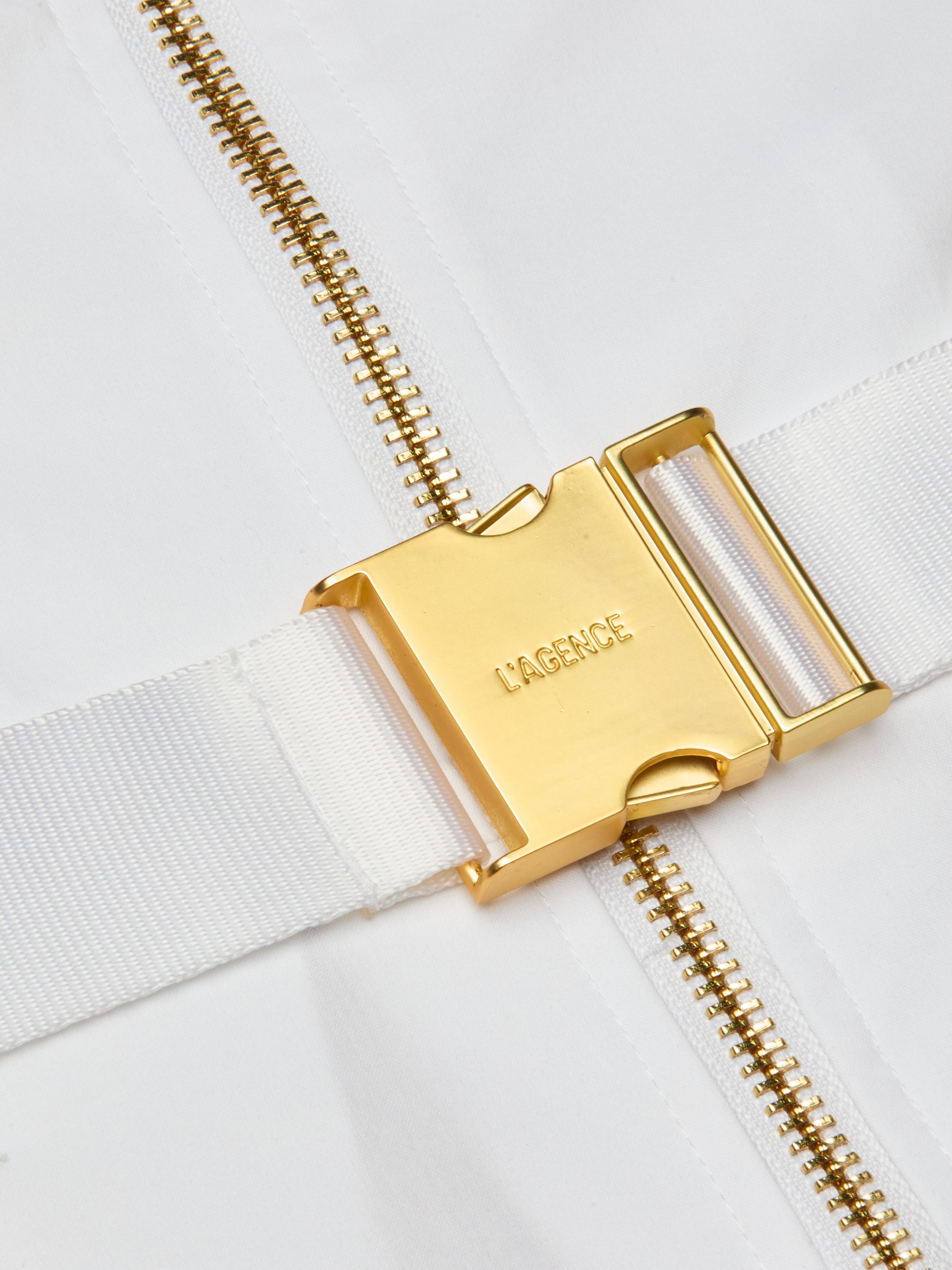 Close-up of a gold metal buckle engraved with LAGENCE on a white fabric strap, next to a gold zipper on a white background, highlighting the refined finish of the Kylo Poplin Shirt Dress crafted from organic cotton.