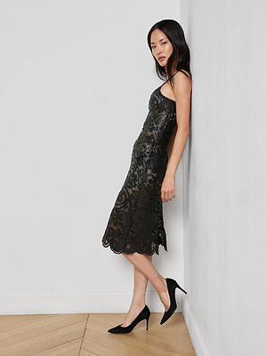 A woman in the Kazuko Vegan Leather Dress, a sleeveless knee-length black midi, stands sideways against a white wall in black heels, looking at the camera with a neutral expression.