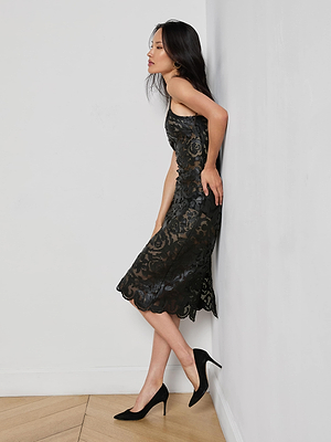 A woman with long dark hair and a calm expression stands sideways, eyes closed, leaning against a white wall in the Kazuko Vegan Leather Dress and black heels. The floor beneath her is light wood.