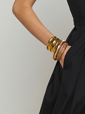 A person wearing a sleeveless black dress has their hand in a pocket, showing several chunky gold and bronze bangles on their wrist against a plain background.