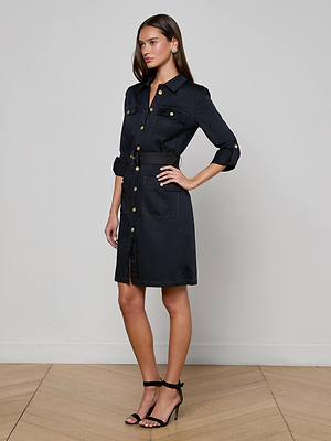 A woman stands sideways in the L'AGENCE Karin Poplin Shirt Dress—dark cotton with rolled-up sleeves, front buttons, and an adjustable belt—paired with black heels, against a light wall and wooden floor.