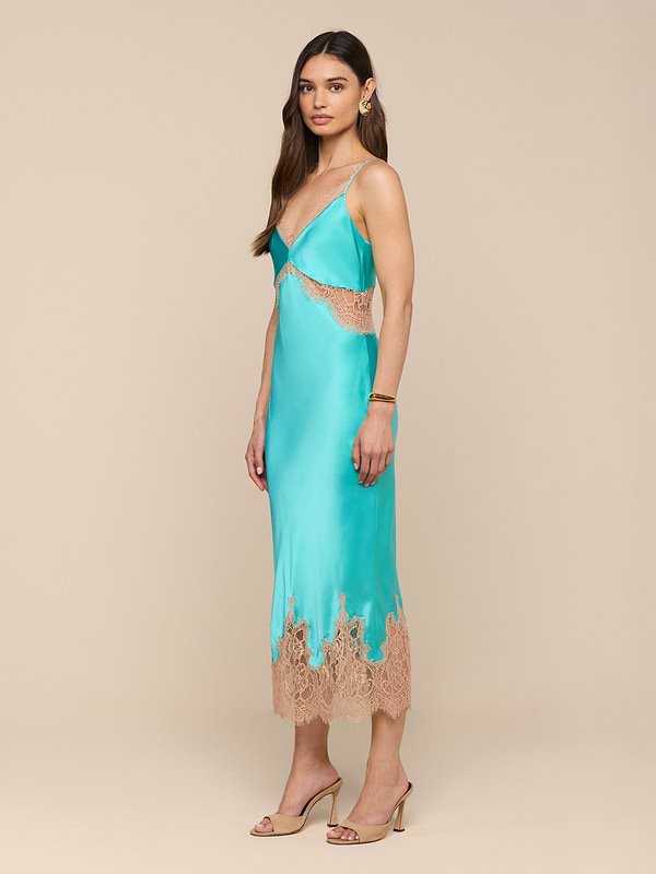 A woman stands against a plain white background wearing the Scotly Silk Lace Dress—a turquoise, bias-cut silk slip dress featuring beige lace trim and lace insets—styled with beige high-heeled sandals and jewelry.
