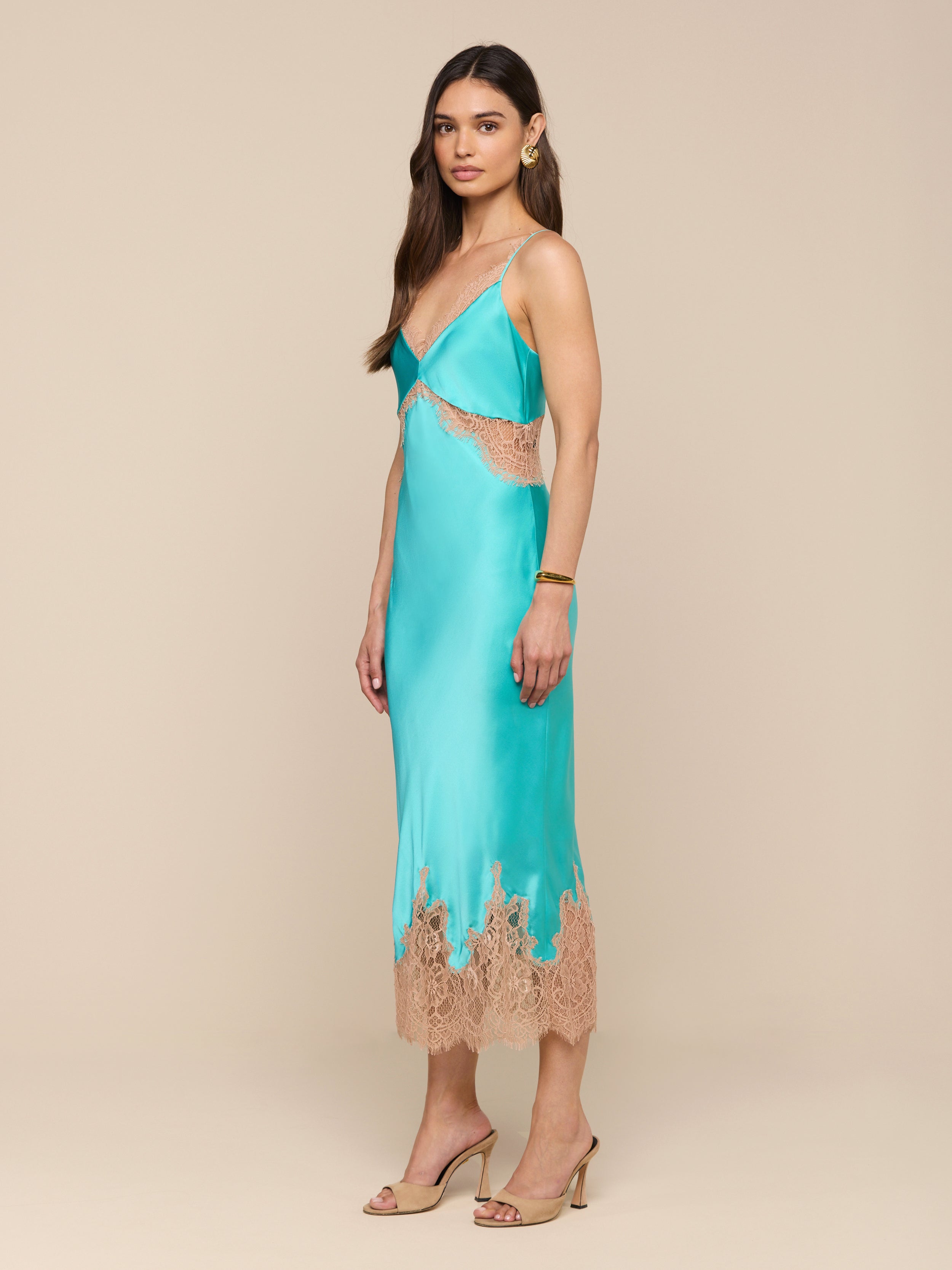 A woman stands against a plain white background wearing the Scotly Silk Lace Dress—a turquoise, bias-cut silk slip dress featuring beige lace trim and lace insets—styled with beige high-heeled sandals and jewelry.
