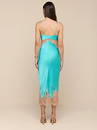 A woman stands facing away against a plain white background, wearing the Scotly Silk Lace Dress—a bias-cut teal silk midi dress featuring lace insets at the hem and back—paired with beige heeled sandals.